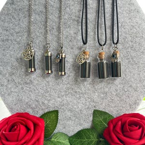 May include: A collection of necklaces featuring small, dark-colored vials and cylindrical pendants. Some pendants have silver chains and charms, including a pentagram and an evil eye. The necklaces are displayed against a gray background with red roses at the bottom.