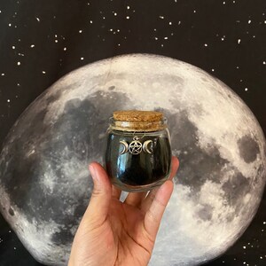 May include: A small glass jar with a cork lid, filled with black material. A silver charm with a triple moon and pentacle design hangs from the jar. The background features a large moon and a starry night sky.