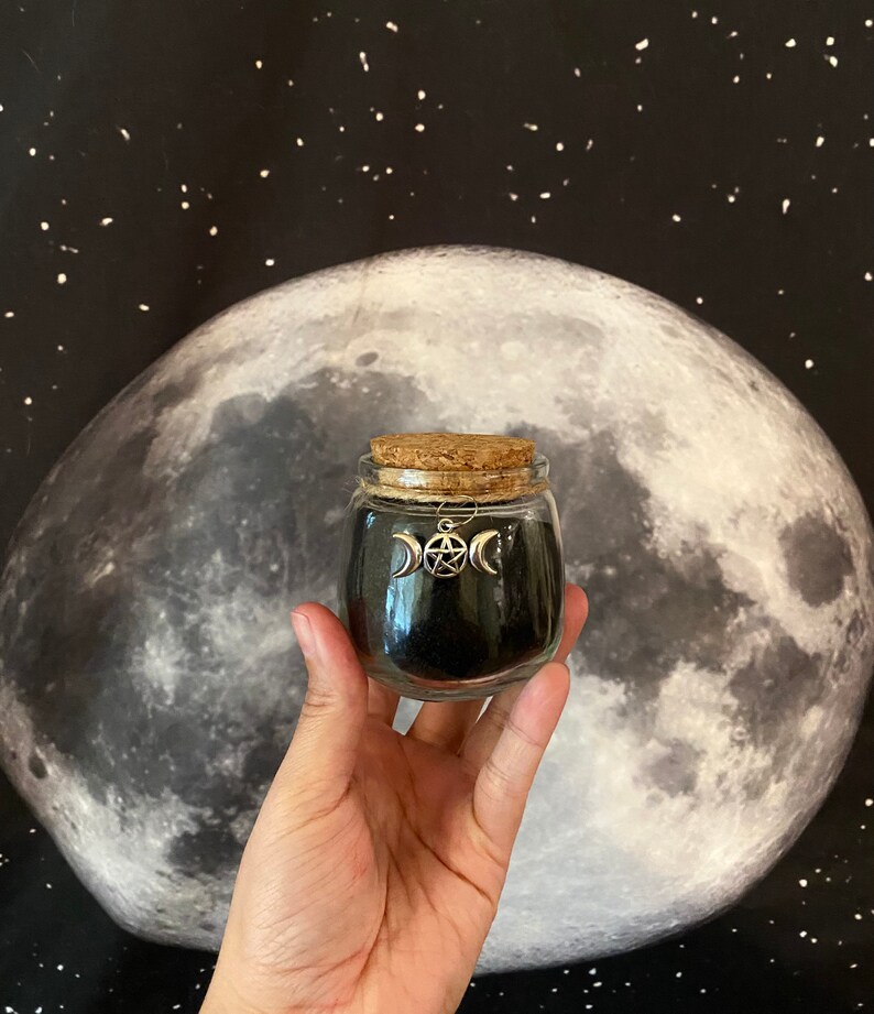 May include: A small glass jar with a cork lid, filled with black material. A silver charm with a triple moon and pentacle design hangs from the jar. The background features a large moon and a starry night sky.