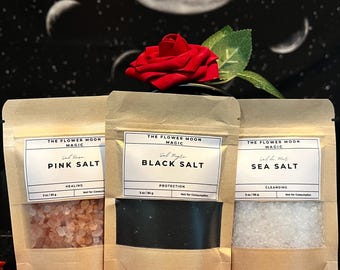 Puede incluir: Tres bolsas de papel kraft etiquetadas como "Pink Salt", "Black Salt" y "Sea Salt" con una rosa roja encima. Cada bolsa tiene una etiqueta blanca con texto negro. El fondo presenta un diseño celestial con una gran luna y estrellas.