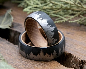 May include: Two black and silver wedding bands with a wood inlay and a forest silhouette design. The rings are on a wooden surface with green leaves.