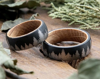 May include: Two black and wood rings with a silver tree design. The rings are on a wooden surface with green foliage.