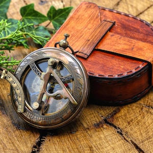May include: A round brass sundial compass with an engraved dial and a folding gnomon, accompanied by a brown leather case with a stitched strap closure. The compass features visible degree markings and directional indicators on its metallic surface.