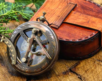 May include: A round brass sundial compass with an engraved dial and a folding gnomon, accompanied by a brown leather case with a stitched strap closure. The compass features visible degree markings and directional indicators on its metallic surface.