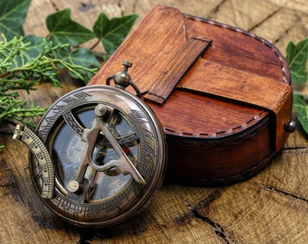 May include: A brass compass with a leather case. The compass has a glass face with a compass rose and a sundial. The case is brown leather with stitching and a metal clasp.