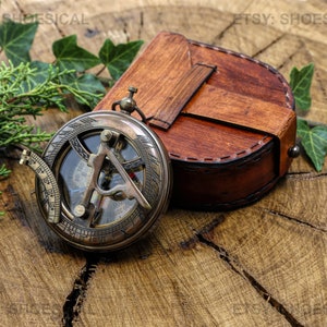 May include: A brass compass with a leather case. The compass has a glass face with a compass rose and a sundial. The case is brown leather with stitching and a metal clasp.
