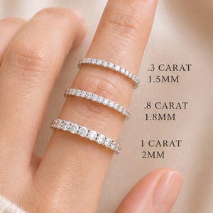 May include: Three diamond eternity rings of varying sizes are displayed on a finger. The rings are labeled with their carat weight and millimeter width: .3 carat 1.5mm, .8 carat 1.8mm, and 1 carat 2mm. The rings are set against a cream-colored background.