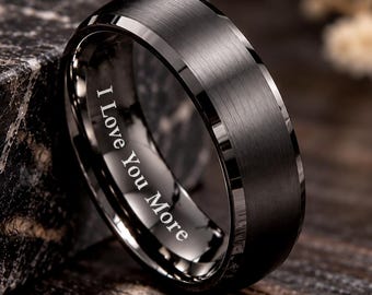 May include: A black ring with a brushed finish and beveled edges. The inner band features the engraved text I Love You More in a clear, white font. The ring has a smooth, metallic appearance with a dark, matte surface texture.
