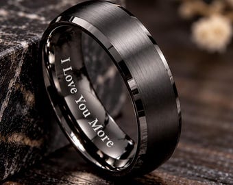 May include: A black ring with a brushed finish and beveled edges. The inner band features the engraved text I Love You More in a clear, white font. The ring has a smooth, metallic appearance with a dark, matte surface texture.