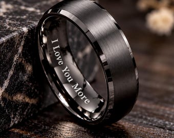 May include: A black ring with a brushed finish and beveled edges. The inner band features the engraved text I Love You More in a clear, white font. The ring has a smooth, metallic appearance with a dark, matte surface texture.