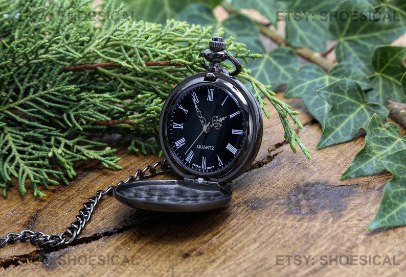 May include: A black pocket watch with Roman numeral hour markers and a black face. The watch has a silver chain and is sitting on a wooden surface with green leaves.