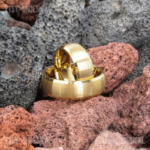 May include: Two gold-colored rings with a brushed matte finish and polished beveled edges. The rings are stacked together, with one positioned upright behind the other, resting against a backdrop of porous black and reddish-brown volcanic rocks.