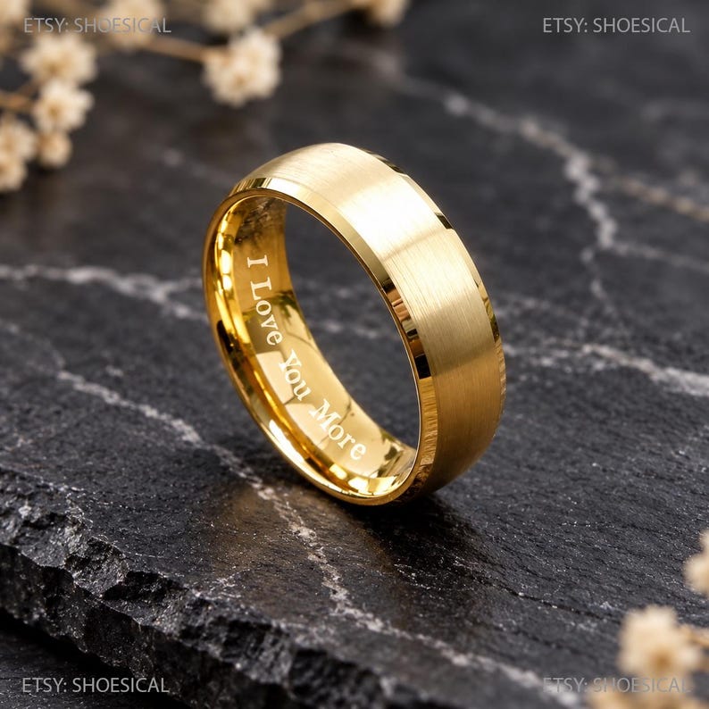 May include: A gold-toned metal ring with a brushed finish and beveled edges. The inner band features the engraved text I Love You More in a clear, sans-serif font. The ring has a polished interior surface and a solid, circular band structure.