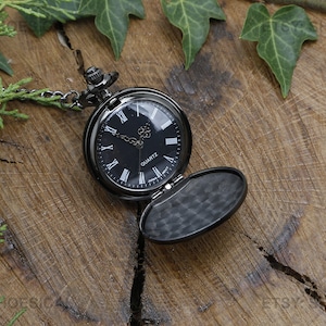 May include: A black pocket watch with a black chain and a black face with Roman numeral markings. The watch is open and the inside is black. The watch is on a wooden surface with green leaves.