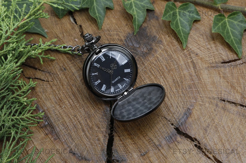 May include: A black pocket watch with a black chain and a black face with Roman numeral markings. The watch is open and the inside is black. The watch is on a wooden surface with green leaves.