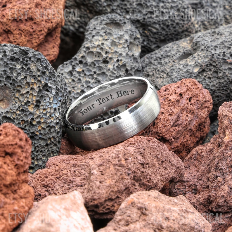Pu&ograve; includere: Una fede nuziale in argento con finitura spazzolata e un'incisione personalizzata che recita "Your Text Here". La fede &egrave; fotografata su un letto di rocce grigio scuro e rosso-marrone.