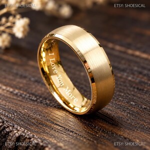 May include: A gold-toned ring with a brushed matte exterior finish and a polished inner band. The interior of the ring features the engraved text I Love You More in a clear, sans-serif font, visible against the reflective gold surface.