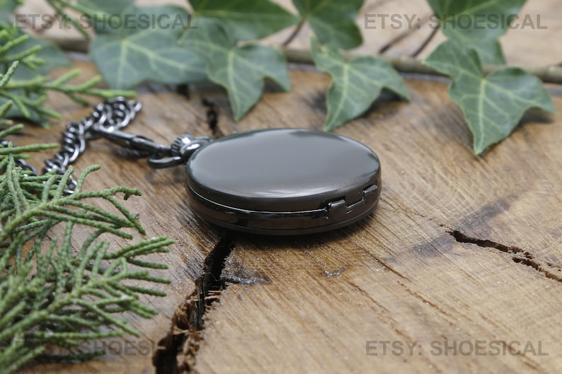 May include: A black pocket watch with a chain on a wooden surface. The watch has a silver finish and a hinged lid.