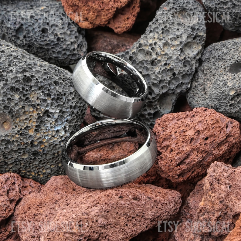 Pu&ograve; includere: Due anelli in carburo di tungsteno argentati con bordi smussati e finitura spazzolata. Gli anelli sono impilati uno sopra l'altro e sono su un letto di rocce grigio scuro e rosse.