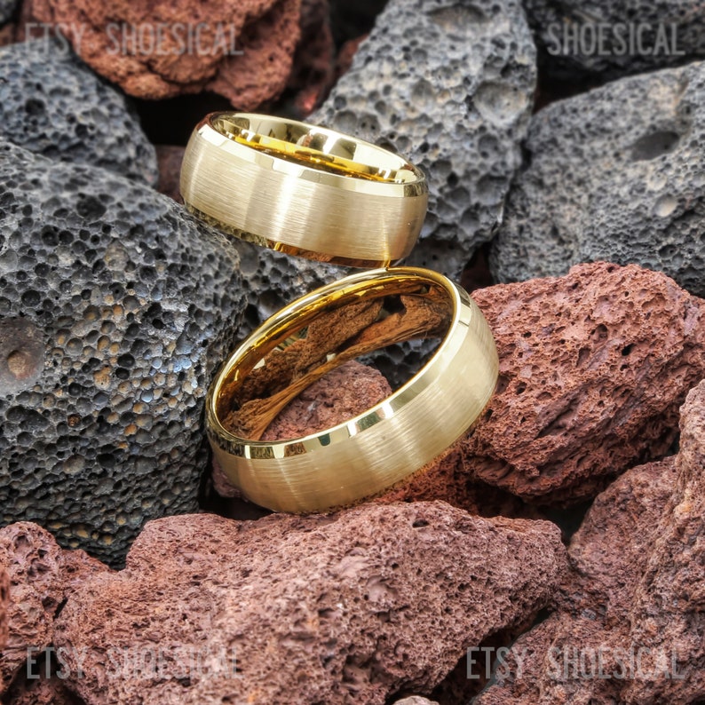 May include: Two gold-toned rings with a brushed matte finish on the exterior and a polished, reflective interior surface. The rings are positioned against a background of porous, dark gray and reddish-brown volcanic rock fragments.