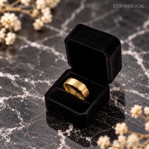 May include: A gold-toned metal ring with a smooth, matte finish resting inside a black velvet jewelry box. The inner band of the ring features the engraved text "I Love You More" in a clear, sans-serif font, centered within the box.