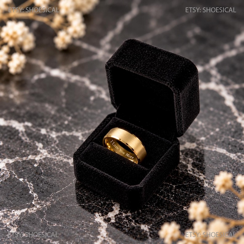 May include: A gold-toned metal ring with a smooth, matte finish resting inside a black velvet jewelry box. The inner band of the ring features the engraved text "I Love You More" in a clear, sans-serif font, centered within the box.