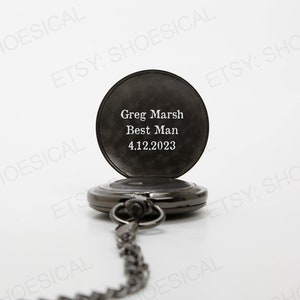 May include: Black pocket watch with a chain. The watch face is engraved with the text "Greg Marsh Best Man 4.12.2023".