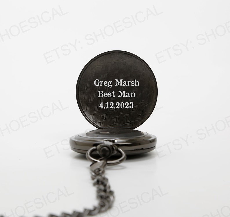 May include: Black pocket watch with a chain. The watch face is engraved with the text "Greg Marsh Best Man 4.12.2023".