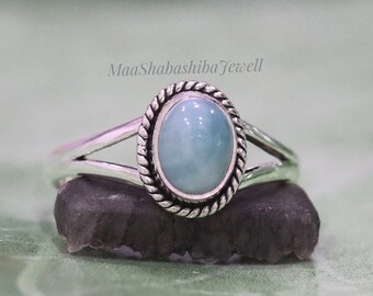 May include: A silver ring featuring an oval, light blue cabochon stone set within a twisted rope-style metal border. The band has a split design on both sides. The text MaaShabashibaJewell is visible above the ring, which rests on a dark stone.