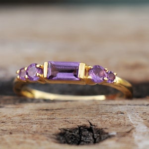 Puede incluir: Un anillo de oro con una piedra de amatista púrpura en el centro y piedras de amatista más pequeñas a cada lado.