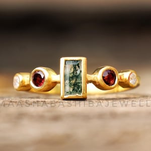 Op de afbeelding: Gouden ring met een rechthoekige groene steen en twee ronde rode stenen. De ring heeft ook kleine witte stenen.