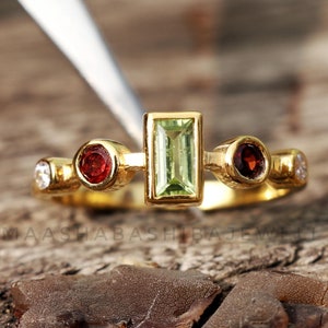 May include: A gold ring featuring a rectangular green gemstone at the center, flanked by two red gemstones and two smaller white gemstones. The ring is set on a wooden surface.
