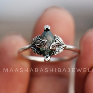 Puede incluir: Anillo de plata con una gema de talla marquesa con inclusiones verdes y marrones, enmarcada por delicadas hojas. El anillo tiene una banda delgada y un diseño de temática botánica.