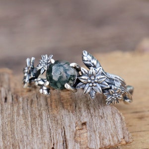 May include: A silver ring featuring a green moss agate stone and floral details. The ring has a delicate design with leaves and flowers.