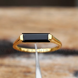 Puede incluir: Un anillo dorado con una piedra negra rectangular. El anillo tiene un diseño simple y elegante, con una banda lisa y un engaste de bisel para la piedra negra. Se muestra sobre una superficie de madera.