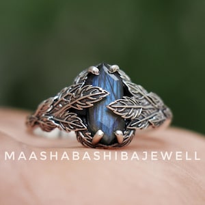 Pode incluir: Um anel de prata com uma grande pedra preciosa azul. A pedra é rodeada por um design detalhado de folhas. O anel está sobre um fundo verde desfocado. O texto "MAASHABASHIBAJEWELL" é visível.