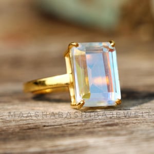 Könnte beinhalten: Ein goldener Ring mit einem großen, klaren, rechteckigen Edelstein. Der Edelstein hat einen regenbogenfarbenen, irisierenden Effekt.