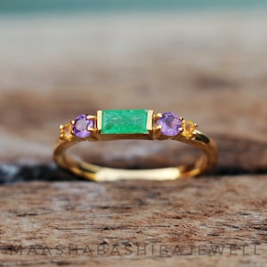 Könnte beinhalten: Ein goldener Ring mit einem grünen Smaragd und violetten Amethyst-Steinen. Der Ring liegt auf einer Holzoberfläche.