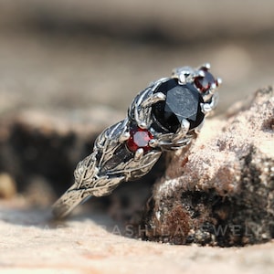Könnte beinhalten: Ein silberner Ring mit einem schwarzen Mittelstein und zwei kleineren roten Steinen. Der Ring hat ein detailliertes Blattdesign auf dem Band und um die Steine. Der Ring ist vor einem verschwommenen, natürlichen Hintergrund platziert.