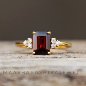 Op de afbeelding: Een gouden ring met een grote, rechthoekige, donkerrode edelsteen omringd door kleinere, heldere edelstenen.