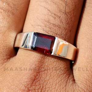 Op de afbeelding: Een zilveren ring met een rechthoekige, donkerrode edelsteen in het midden.