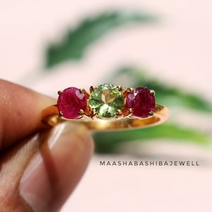 May include: A gold ring set with three gemstones. The central stone is a light green peridot, and the two stones on either side are red rubies.