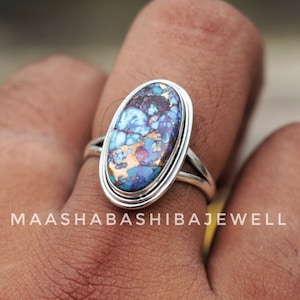May include: Oval-shaped silver ring with a large, colorful stone. The stone features a mix of blue, purple, and copper hues. The ring has a double-band design and the text "MAASHABASHIBIBAJEWELL" is visible.
