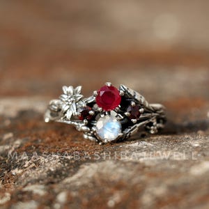 Könnte beinhalten: Ein silberner Ring mit floralem und Blättermuster, mit einem großen, runden, roten Edelstein und einem kleineren, runden, weißen Edelstein. Zusätzliche kleinere rote Edelsteine sind im Design des Rings gesetzt. Der Ring liegt auf einer strukturierten Oberfläche.