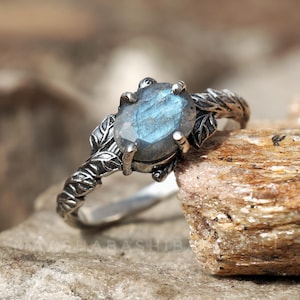 Könnte beinhalten: Ein silberner Ring mit einem ovalen, blau-grauen Edelstein. Der Ring hat ein detailliertes Blattdesign am Band und um die Edelsteinfassung. Der Edelstein hat ein poliertes, facettiertes Aussehen.