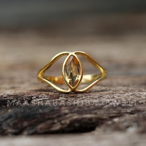 Peut inclure: Une bague dorée avec une pierre précieuse centrale ovale de couleur marron clair. La monture de la bague se divise en deux sections incurvées qui maintiennent la pierre. La bague est posée sur une surface en bois rustique.