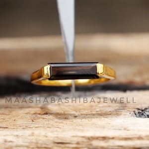 Op de afbeelding: Een gouden ring met een donkerbruine rechthoekige edelsteen. De ring ligt op een houten oppervlak.