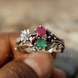 Puede incluir: Un anillo de plata con un diseño floral, con una flor y hojas. El anillo está adornado con una gema roja grande y redonda, una gema roja más pequeña y una gema verde. El texto "MAASHABASHIBAJEWELL" es visible.