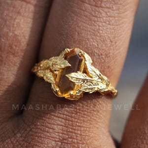 May include: A gold ring with an amber-coloured, rectangular gemstone. The ring has a leaf design on the band. The text "MAASHABASH JEWELL" is visible on the ring.