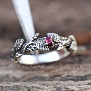 Puede incluir: Un anillo de plata con un delicado diseño de rama y una pequeña piedra preciosa rosa. El anillo está colocado sobre una superficie de madera.